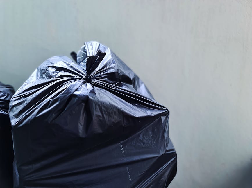 Close-up view of several black plastic rubbish bags filled with waste, stacked against a plain, light-colored wall in an outdoor or garage setting. The bags are tightly tied at the top, with the mater